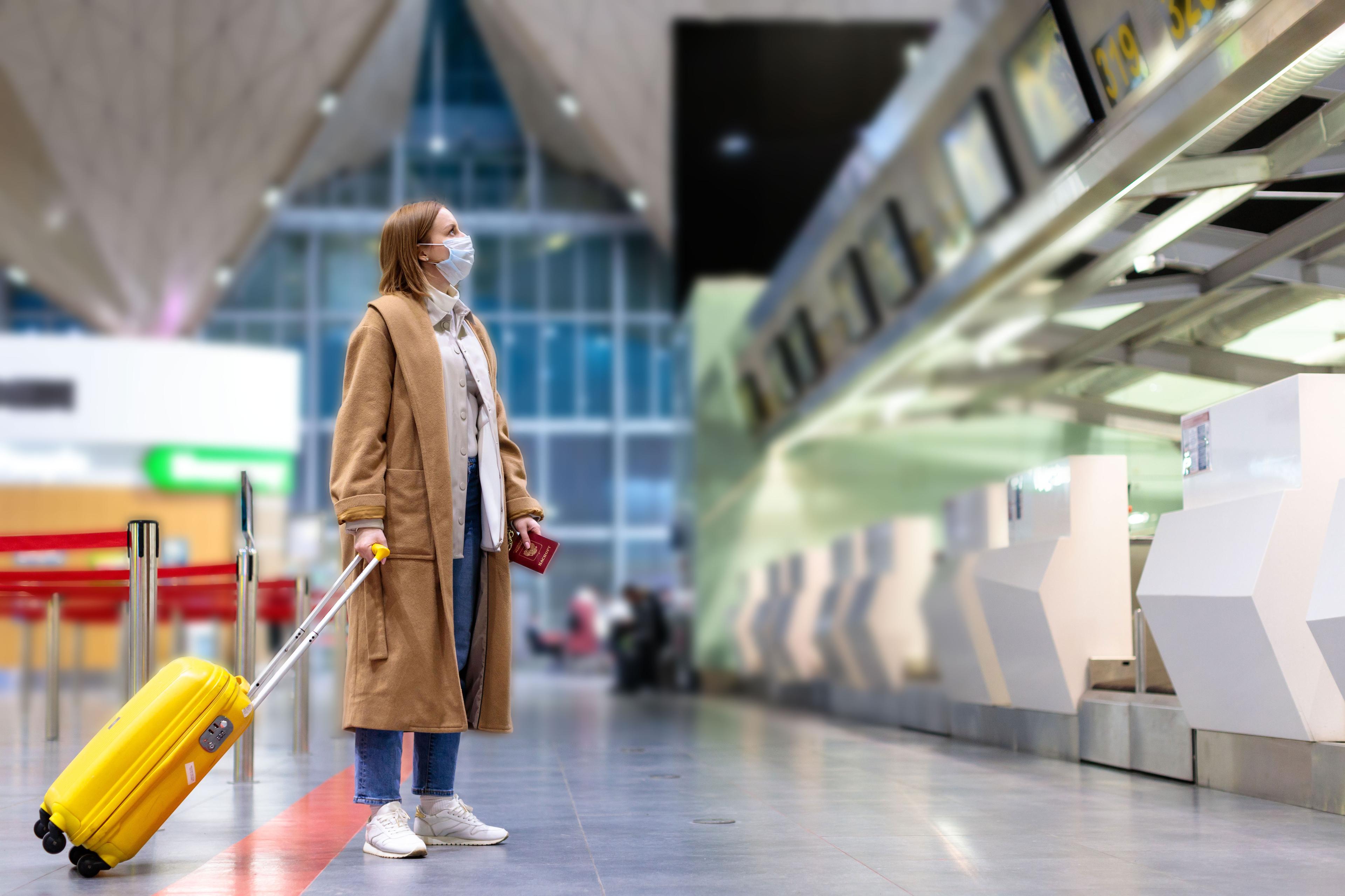 A woman standing with her luggage at an airport, wearing a face mask